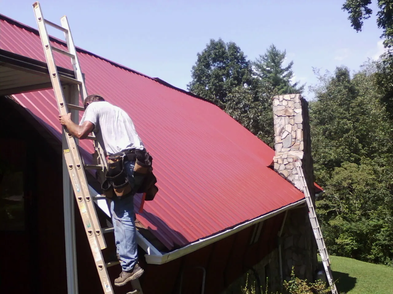 Roofer on a ladder beside a red metal roof and stone chimney — Soffit & Fascia Repair by Roofing Brothers in Mount Ulla
